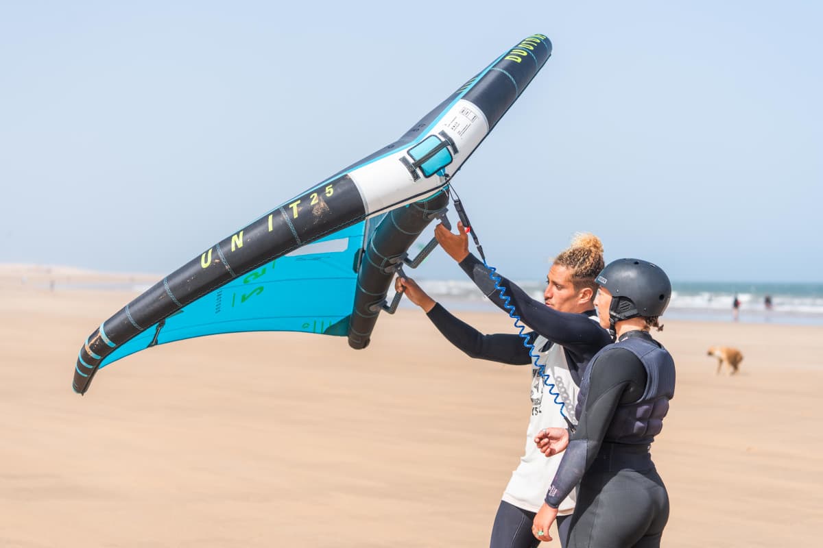 A wingfoiler gliding silently above the water surface on a foil. The wing is held high, and the board is elevated above the waves. The lighting is high-key and bright, emphasizing the sensation of flight. Minimalist ocean background with deep blue tones. High-energy technical watersport aesthetic.